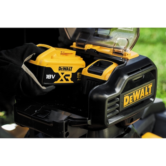 A close-up of a person wearing work gloves inserting a DEWALT 18V XR lithium-ion battery into a DEWALT power tool or equipment. The battery compartment is open, revealing the slot for the battery, and the DEWALT logo is visible on the device.