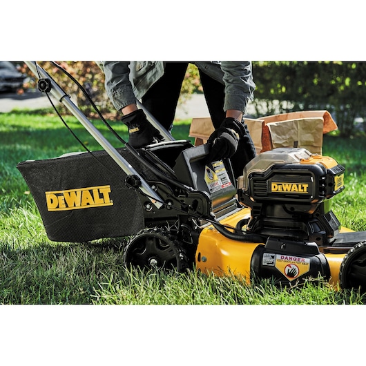 Close-up of a person operating a DEWALT lawn mower on green grass. The person is wearing gloves and handling the mower's compartment. The DEWALT logo is clearly visible on the grass bag and the mower body.