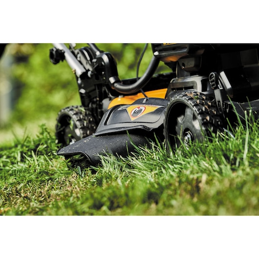 Close-up view of a DEWALT cordless lawn mower cutting grass, focusing on the side discharge chute and rear wheel on a green lawn.