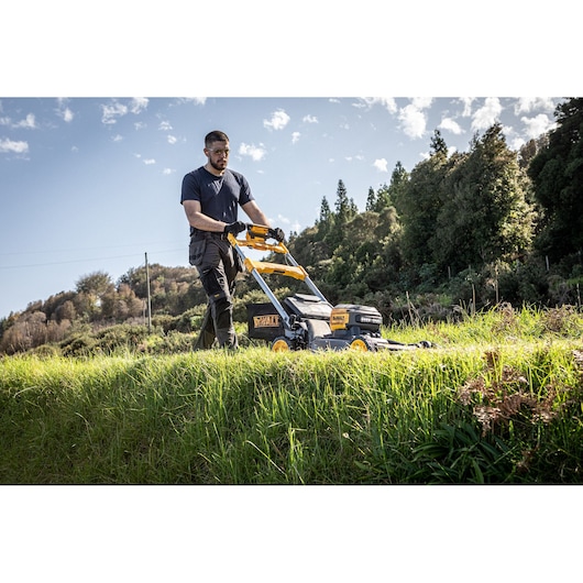 A person uses a DEWALT cordless lawn mower (model DCMWSP660N) outdoors on grassy terrain with trees in the background. The