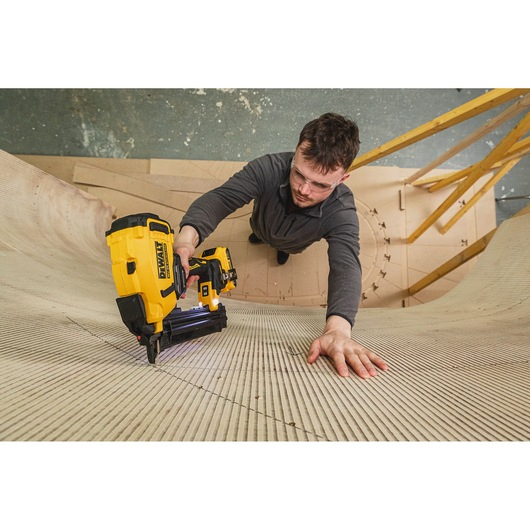 Person uses a DEWALT cordless nailer to fasten curved wood panels, wearing safety glasses in a workshop.
