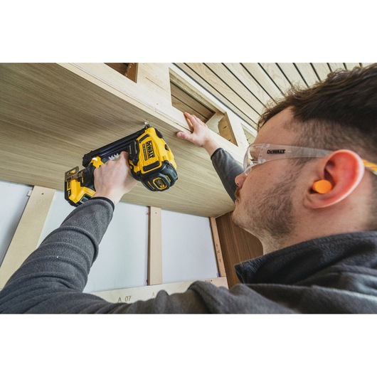Person uses DEWALT nailer to attach wooden panel, wearing safety glasses and earplugs during construction.