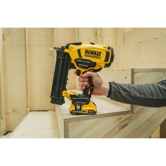 Hand holding a DEWALT cordless nail gun, positioned on a wooden staircase at a construction site.