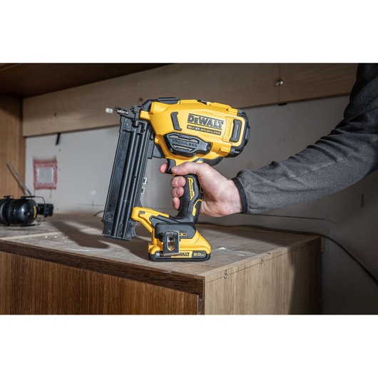 Hand holding a DEWALT cordless nail gun above a wooden cabinet in a workshop setting.
