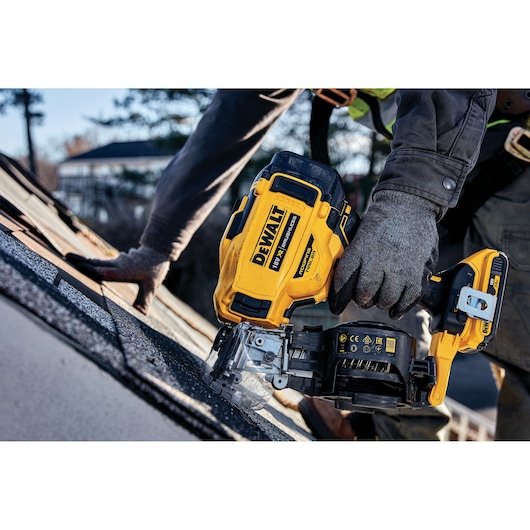 A person wearing work gloves uses a DEWALT cordless roofing nailer on a shingled roof.