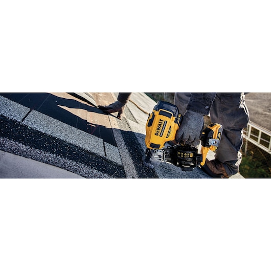 A person wearing gloves and work clothes is using a DEWALT DCN45RND2 roofing nailer to install shingles on a roof.