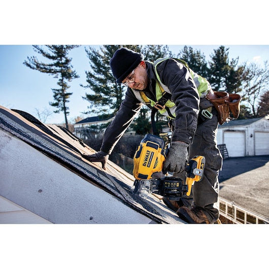 A construction worker using a DEWALT DCN45RND2 cordless roofing nailer while installing shingles on a sloped roof. The