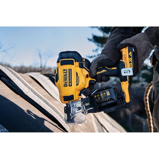 A person wearing gloves is using a DEWALT cordless roofing nailer on a shingled roof outdoors.