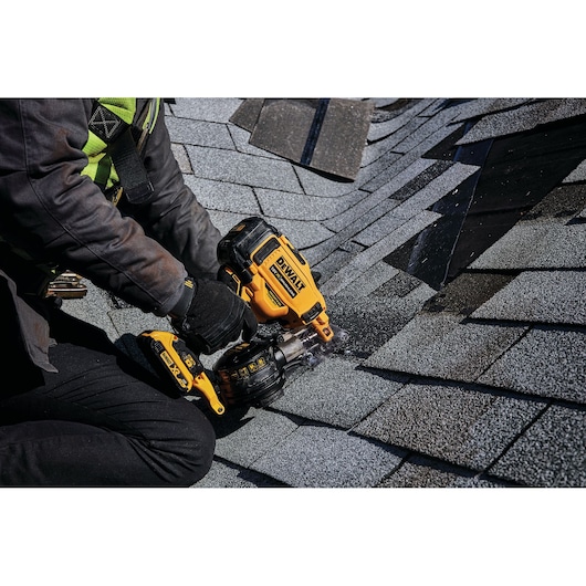 A person using a DEWALT cordless roofing nailer to install shingles on a roof. The tool is yellow and black, and the person