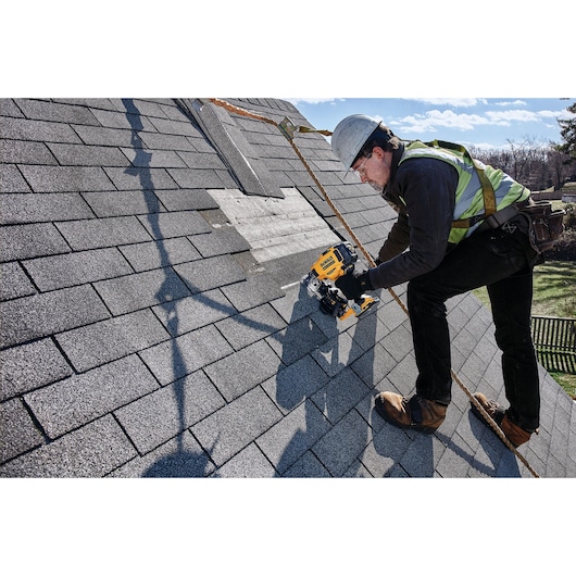 A worker wearing safety gear uses a DEWALT roofing nailer to install shingles on a sloped roof.