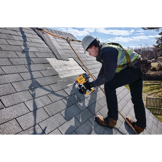 A person wearing safety gear uses a DEWALT DCN45RND2 nailer to install shingles on a roof. The person is secured with a