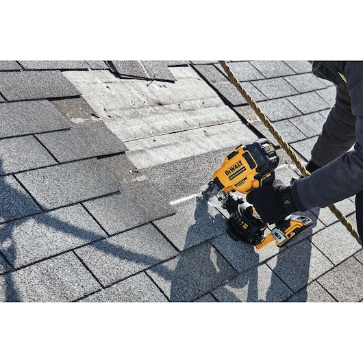 A person using a DEWALT cordless nailer to install roofing shingles on a roof.