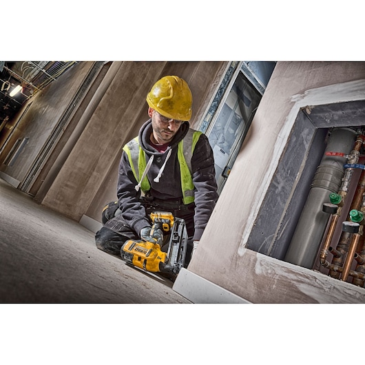 A worker wearing a yellow hard hat and safety vest kneels on the floor, using a DEWALT DCN660D2 cordless nailer to install