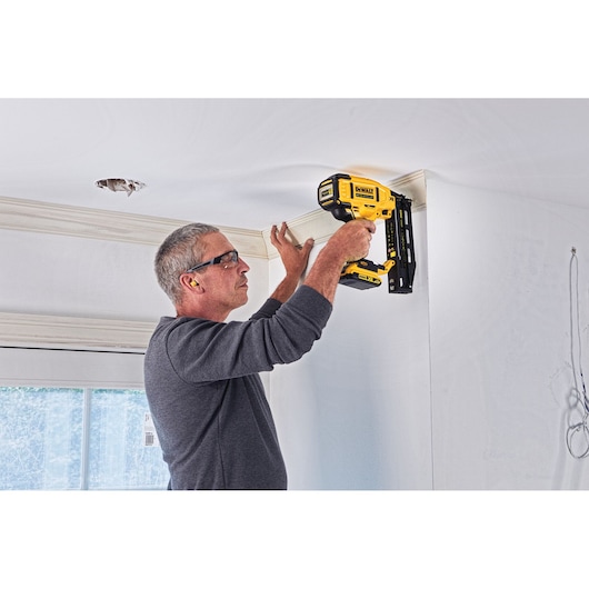A person using a DEWALT DCN662D2 cordless nailer to install trim along a ceiling edge in a room. The individual is wearing