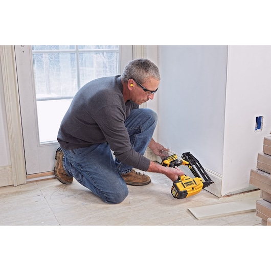 A person kneeling and using a yellow DEWALT cordless nailer (model DCN662D2) to install trim along the base of a wall in a