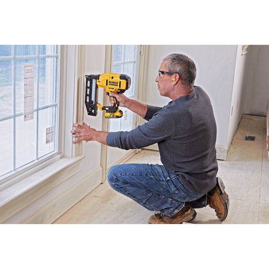 A person kneeling and using a DEWALT DCN662N cordless nailer to install trim on a window frame inside a room. The individual