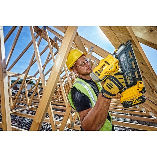 A construction worker wearing a yellow hard hat and safety vest is holding a DEWALT DCN692N nail gun, working on a wooden roof frame structure. The worker's face is blurred for privacy.