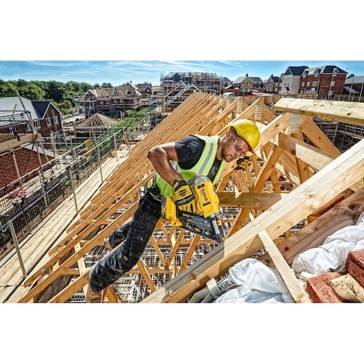 A construction worker wearing a yellow hard hat and a high-visibility vest uses a DEWALT DCN692N nail gun to secure wooden beams on a building site with houses and scaffolding in the background.