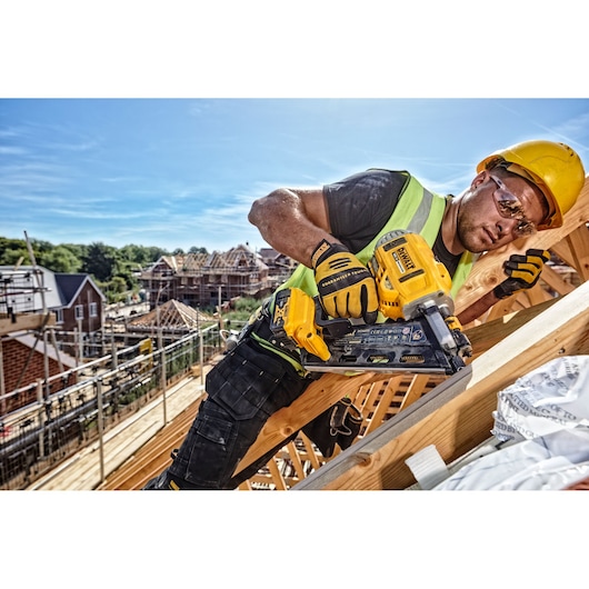 A construction worker wearing a yellow helmet, safety vest, and gloves is using a DEWALT DCN692N cordless nail gun to fasten wooden beams at a building site. Several houses are under construction in the background.