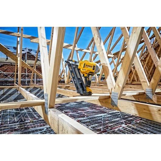 A DEWALT cordless nail gun (model DCN692N) positioned on wooden framing at a construction site, with the tool resting against timber trusses under a clear blue sky.