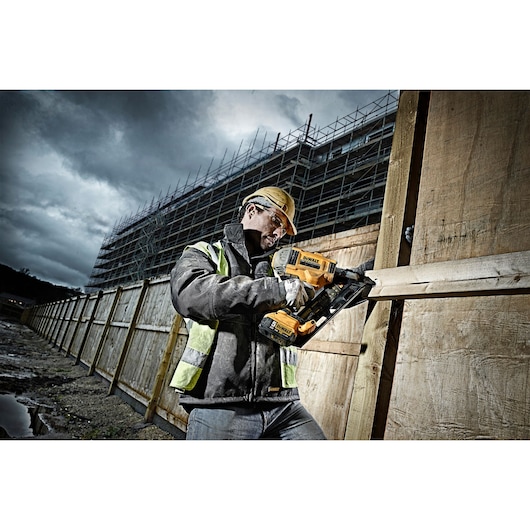 A construction worker in a safety jacket and hard hat using a DEWALT DCN692P2 cordless nailer to attach wooden boards at a building site, with scaffolding and cloudy skies in the background.
