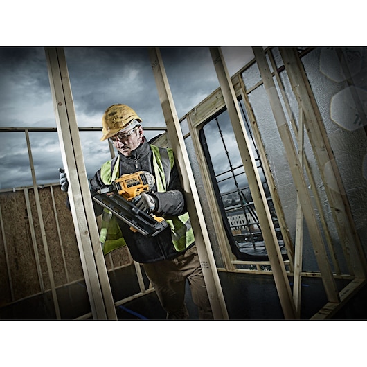 A construction worker wearing a yellow hard hat and high-visibility vest uses a DEWALT DCN692P2 nail gun to fasten wooden frames at a building site. The worker's face is blurred for privacy, and the background shows a partially constructed structure under a cloudy sky.