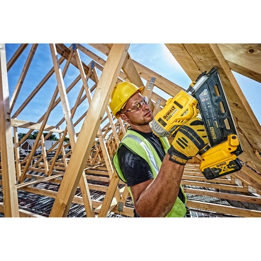 A construction worker wearing a yellow hard hat and safety vest is using a DEWALT nail gun to fasten wooden beams at a building site. The worker is holding the tool confidently, surrounded by wooden framework under a clear blue sky.