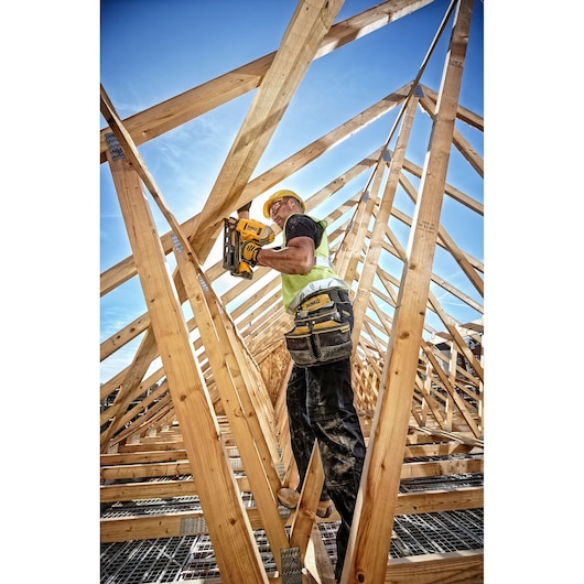 A construction worker wearing a yellow hard hat and safety vest uses a DEWALT nail gun to secure wooden beams on a building roof structure. The worker stands amidst a wooden framework under a clear blue sky.