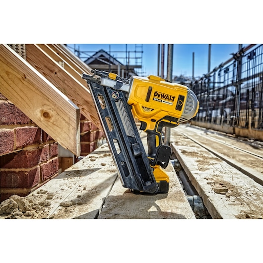 A DEWALT DCN692P2 18V cordless nail gun shown upright on a construction site, with wooden beams and brickwork in the background.