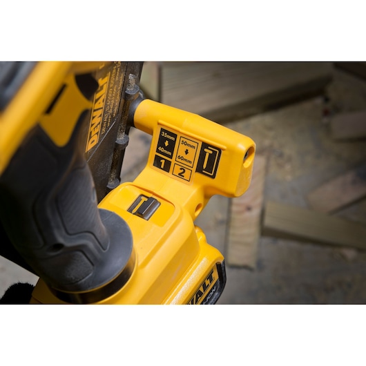 Close-up view of a DEWALT power tool showing its adjustment settings, including options for 35mm, 40mm, 50mm, and 60mm, with a yellow and black color scheme. The tool is being used in a workshop environment with wood pieces in the background.