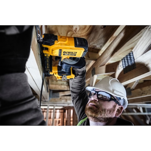 A person wearing protective gear is using a DEWALT 18V XR electric tool to secure cables or wiring to wooden beams in a