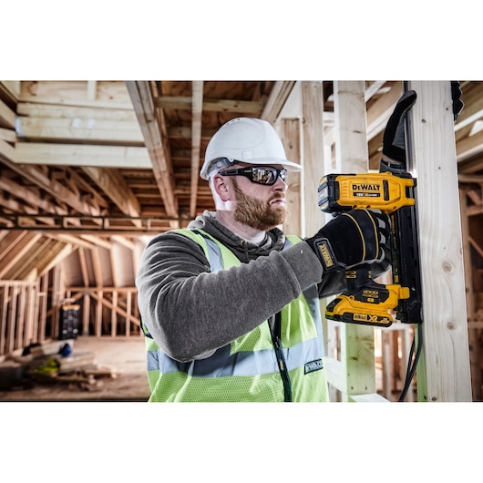 A construction worker wearing safety gear uses a DEWALT DCN701D2 cordless tool to fasten cable clips to a wooden frame