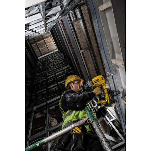 A construction worker wearing a yellow hard hat and a high-visibility vest uses a DEWALT DCN890P2 tool to fasten metal