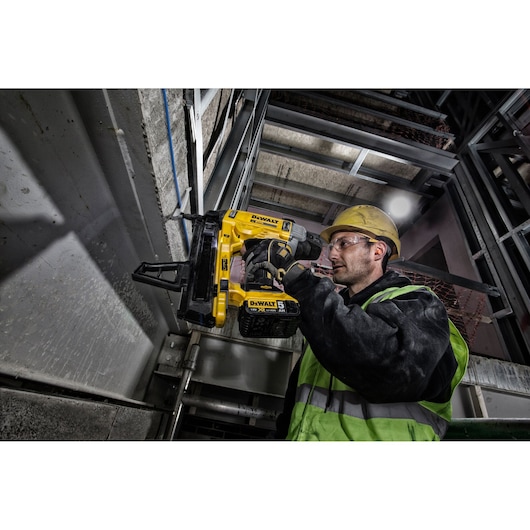 A worker wearing a safety helmet and high-visibility vest is using a DEWALT DCN890P2 tool to fasten objects onto a concrete