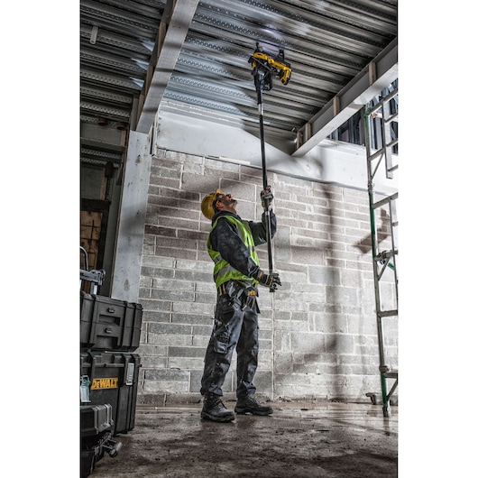 A construction worker wearing a yellow safety helmet and high-visibility vest uses a DEWALT tool attached to an extension