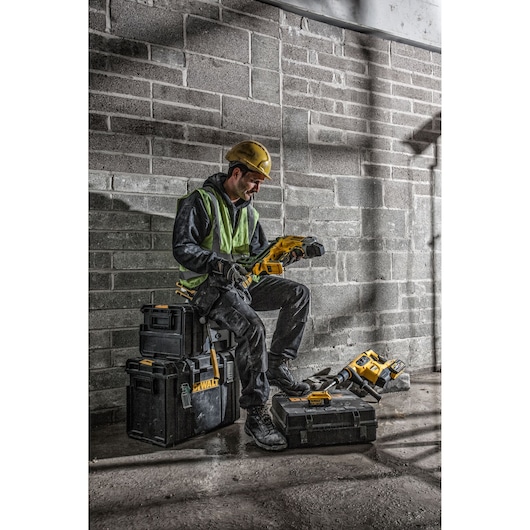 A construction worker wearing a yellow hard hat and a safety vest sits on DEWALT tool storage cases in a rough industrial