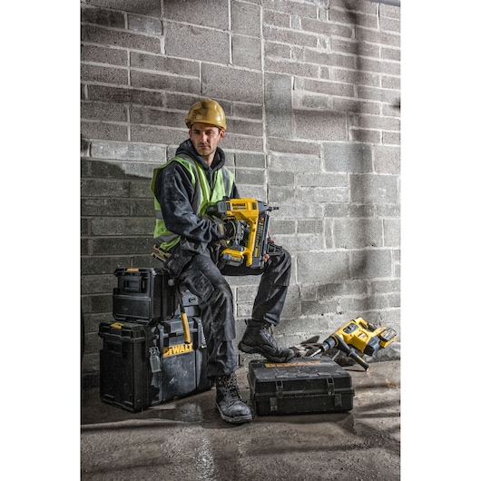 A construction worker wearing a yellow hard hat and safety vest sits on stacked DEWALT toolboxes, holding a DEWALT DCN890P2
