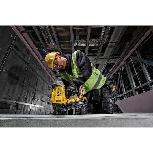 A construction worker wearing a yellow hard hat and green safety vest uses a DEWALT DCN890P2 cordless tool to fasten