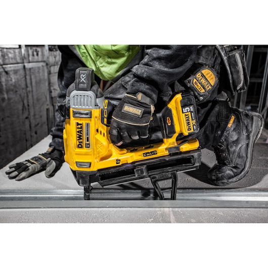 A person wearing protective workwear uses a yellow DEWALT DCN890P2 cordless nailer to secure metal framing. The tool has