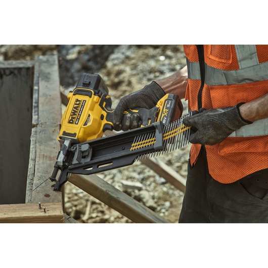 Person uses DEWALT DCN910 nailer to fasten wood outdoors, wearing gloves and safety vest.