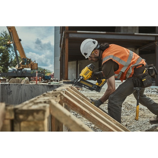 Worker uses DEWALT DCN910 nailer on wood at construction site, wearing safety helmet and orange vest.