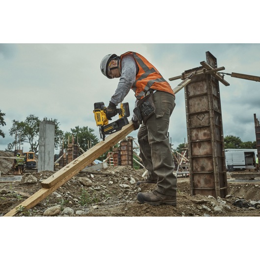 Worker using DEWALT DCN910 nailer on wood at a construction site, wearing safety gear and orange vest.