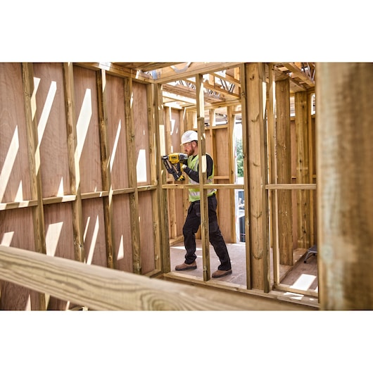 A person wearing a safety vest and helmet uses a DEWALT nailer, model DCN930N, to fasten wooden framing inside a partially constructed building.