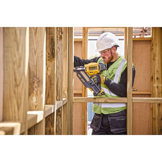 A construction worker wearing a safety vest and helmet is using a DEWALT DCN930N cordless nailer to work on a wooden framework.