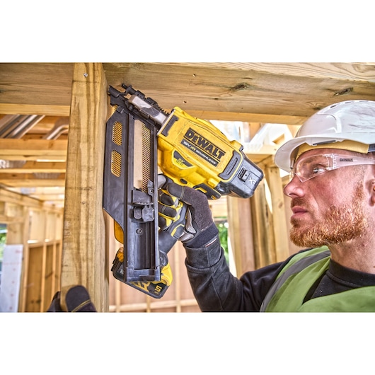 A person wearing a safety helmet and gloves uses a DEWALT DCN930N cordless nail gun to fasten a wooden beam during construction. The face of the individual is blurred.