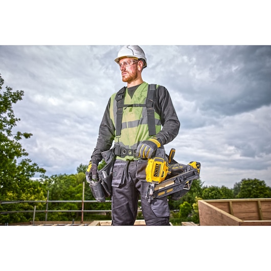 A construction worker with a blurred face is standing outdoors, wearing a safety vest, gloves, and tool belt. The worker is holding a DEWALT DCN930N nailer. The background shows a partially constructed wooden structure and greenery under a cloudy sky.