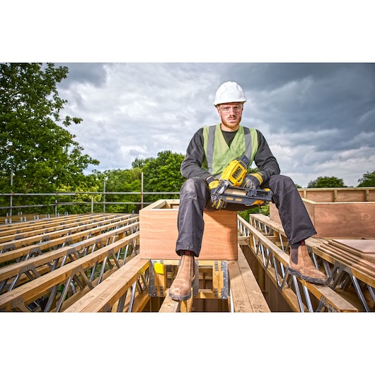 A construction worker wearing a white hard hat, green safety vest, and work boots is sitting on wooden beams at a construction site. The worker is holding a DEWALT DCN930N tool and is surrounded by wooden and metal framework. Trees and cloudy sky are visible in the background.