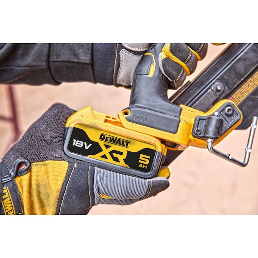 Close-up of a person wearing gloves holding a DEWALT 18V XR 5Ah cordless tool, showing the battery attached to the device.