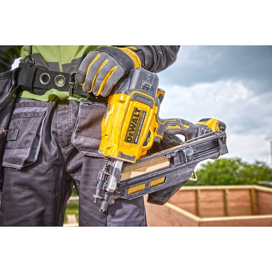 Close-up of a person wearing work gloves holding a DEWALT DCN930N nailer at a construction site, with a wooden structure in the background.