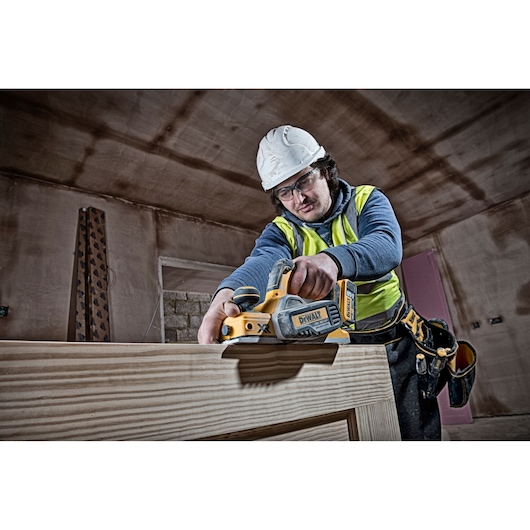 A construction worker wearing a hard hat and safety vest is using a DEWALT DCP580N cordless planer on a wooden surface inside a building under construction.